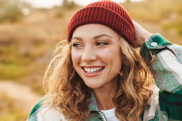 Retrato de uma jovem alegre, caucasiana, usando chapéu e camisa xadrez, sorrindo enquanto caminha ao ar livre