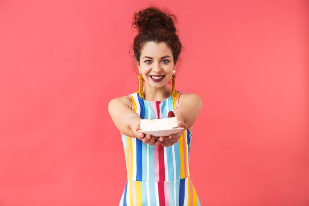 Foto retrato de uma jovem alegre com um vestido de pé isolado sobre um fundo vermelho, segurando o prato com um pedaço de bolo