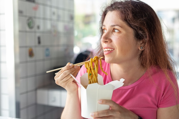 Retrato de uma jovem alegre comendo macarrão chinês em um café e olhando pela janela. O conceito de cozinha asiática saudável.