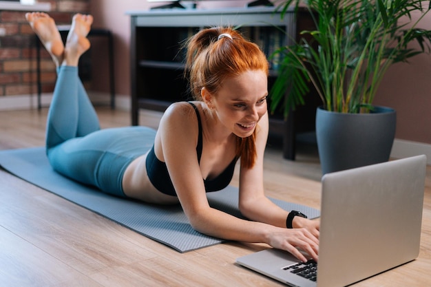 Retrato de uma jovem alegre deitada no chão e usando o computador portátil no apartamento. Mulher sorridente esportiva conversando on-line via notebook enquanto descansa após o treinamento esportivo.