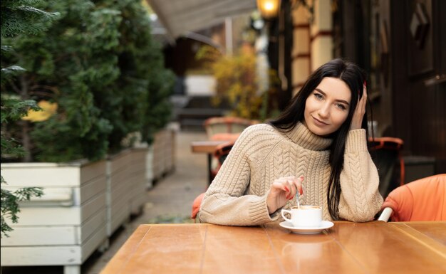 Foto retrato de uma jovem alegre desfrutando de uma xícara de café em um café sorrindo menina bonita bebendo bebida quente no outono mulher excitada em um suéter em um dia de outono