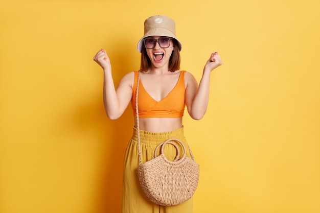 Retrato de uma jovem alegre extremamente feliz vestindo roupas de verão com saco de palha em pé isolado sobre fundo amarelo punhos cerrados ganhando gritando de felicidade