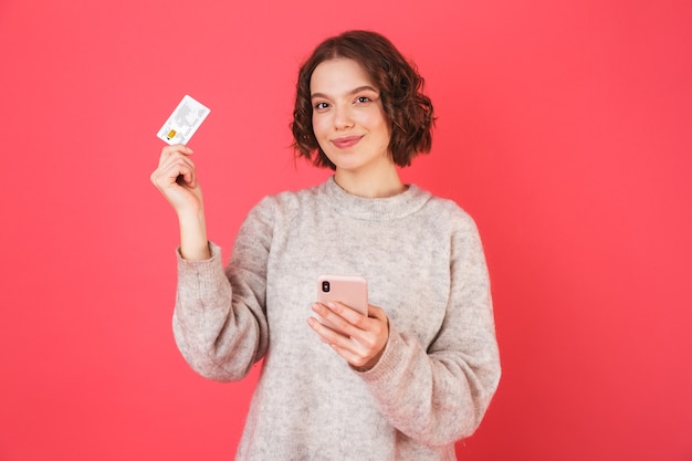 Retrato de uma jovem alegre, isolada sobre rosa, usando um telefone celular, mostrando um cartão de crédito de plástico