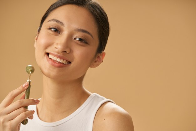 Retrato de uma jovem alegre sorrindo para a câmera, usando o rolo de jade para massagear o rosto