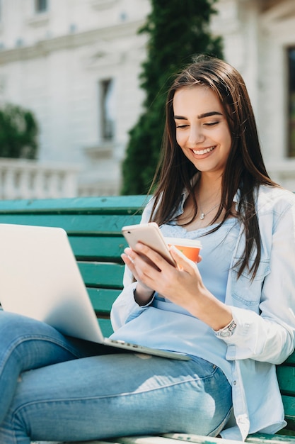 Retrato de uma jovem aluna encantadora sentado em um banco contra a escola, olhando para um smartphone sorrindo, segurando um laptop nas pernas.