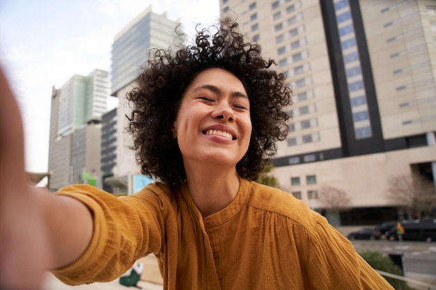 Foto retrato de uma jovem americana sorridente com os olhos fechados e afro tirando uma selfie do lado de fora de um shopping