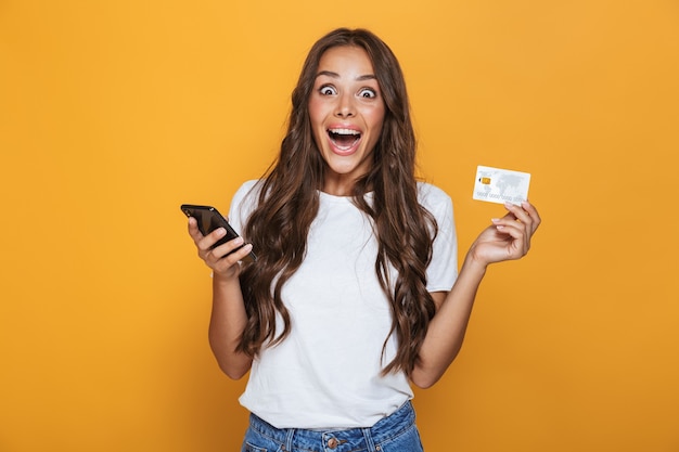 Retrato de uma jovem animada com longos cabelos castanhos em pé sobre uma parede amarela, segurando um telefone celular, mostrando um cartão de crédito de plástico