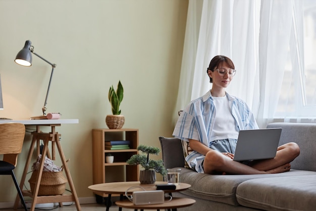 Retrato de uma jovem aproveitando o fim de semana em casa e usando laptop sentado no sofá no interior aconchegante c