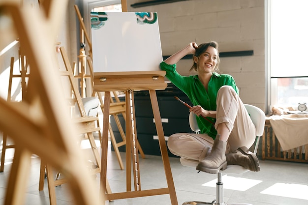 Retrato de uma jovem artista sorridente e muito feliz segurando o pincel no nariz posando para a câmera nas proximidades da tela