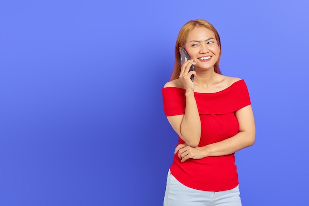 Retrato de uma jovem asiática alegre de vestido vermelho regozijando-se enquanto falava no celular isolado em fundo roxo