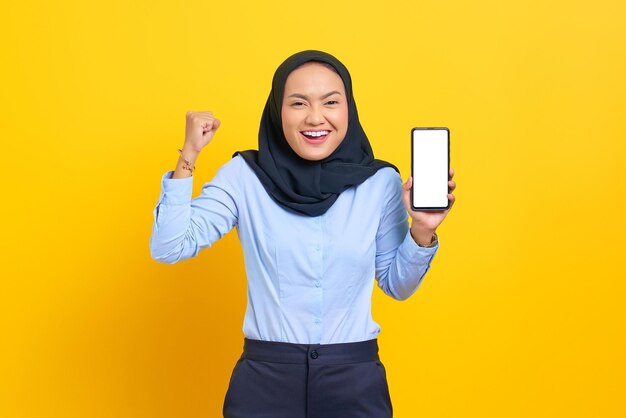 Retrato de uma jovem asiática alegre mostrando um celular de tela em branco e fazendo um gesto de sucesso isolado em um fundo amarelo