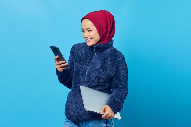 Retrato de uma jovem asiática alegre usando smartphone enquanto segura o laptop nas mãos