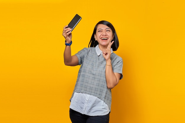 Retrato de uma jovem asiática alegre usando telefone celular, tirar selfie e fazer o sinal da paz