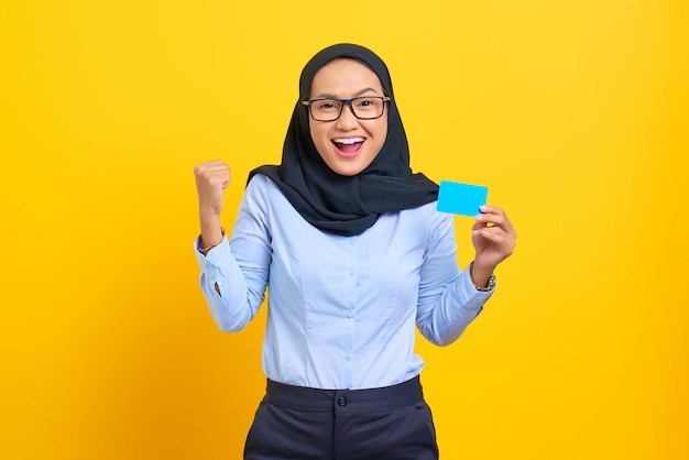 Retrato de uma jovem asiática animada segurando um cartão em branco e levantando o punho em um gesto de sucesso isolado em fundo amarelo