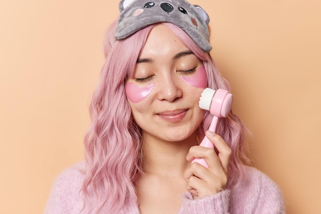 Retrato de uma jovem asiática de cabelo rosa satisfeito usa escova de rosto tem pele limpa e perfeita e saudável aplica adesivos de hidrogel para rejuvenescimento mantém os olhos fechados usa venda e poses de jumper no interior