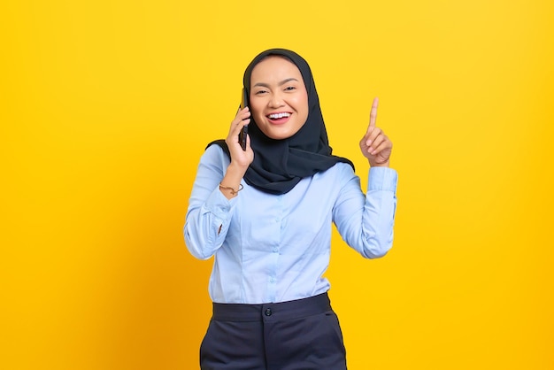 Retrato de uma jovem asiática feliz falando no celular e tendo uma ideia enquanto aponta o dedo isolado no fundo amarelo