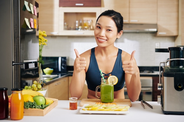 Retrato de uma jovem asiática feliz mostrando os polegares para cima no balcão da cozinha com suco verde que ela fez