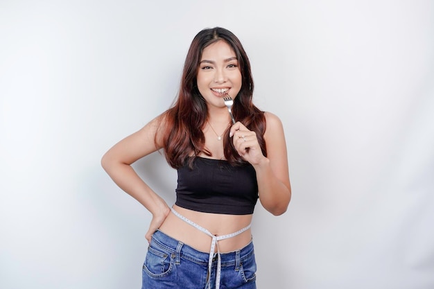 Retrato de uma jovem asiática feliz usando fita métrica ao redor da cintura segurando colher e garfo isolado sobre fundo branco