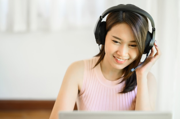Retrato de uma jovem asiática feliz usando um fone de ouvido enquanto trabalha em um notebook em casa