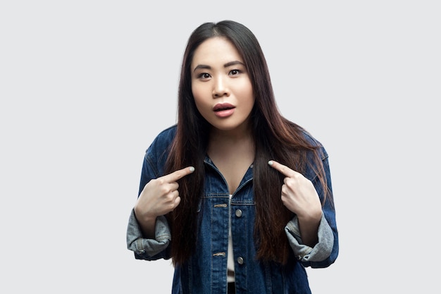 Retrato de uma jovem asiática morena linda chocada em uma jaqueta jeans azul casual com maquiagem em pé, apontando a si mesma e olhando para a câmera. tiro de estúdio interno, isolado em fundo cinza claro.