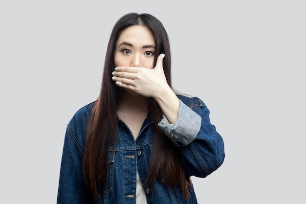 Retrato de uma jovem asiática morena linda chocada na jaqueta azul com maquiagem em pé, cobrindo a boca e olhando para a câmera com olhos grandes. tiro do estúdio, isolado no fundo cinza claro.