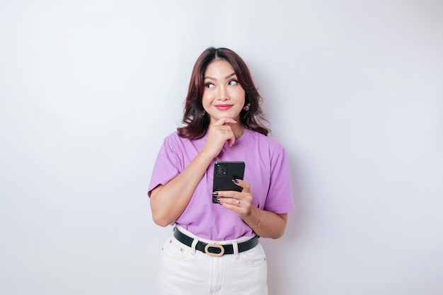 Retrato de uma jovem asiática pensativa vestindo camiseta roxa lilás olhando de lado enquanto segura smartphone