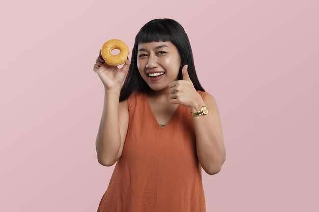 Retrato de uma jovem asiática segurando donuts com os polegares para cima do dedo, isolado sobre fundo rosa