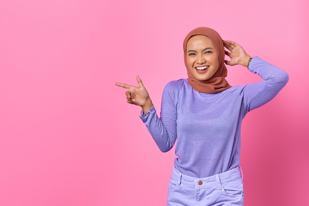 Retrato de uma jovem asiática sorridente apontando o dedo para o espaço da cópia em fundo rosa