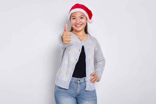 Retrato de uma jovem asiática sorridente com chapéu de Papai Noel aparecendo os polegares isolados sobre um fundo branco