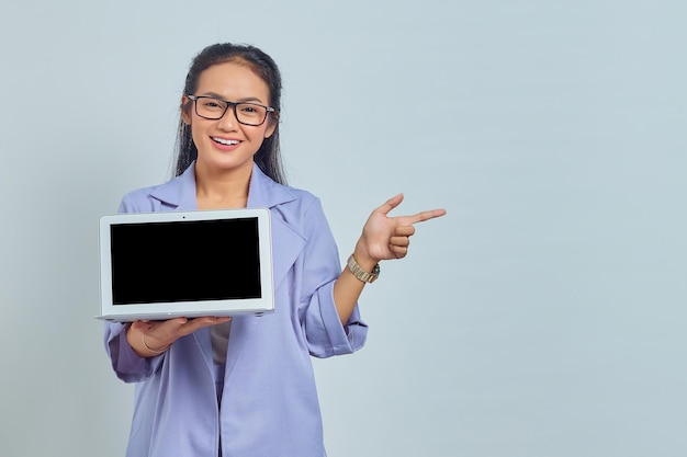 Retrato de uma jovem asiática sorridente mostrando a tela do laptop em branco e apontando para copiar o espaço com o dedo isolado no fundo branco