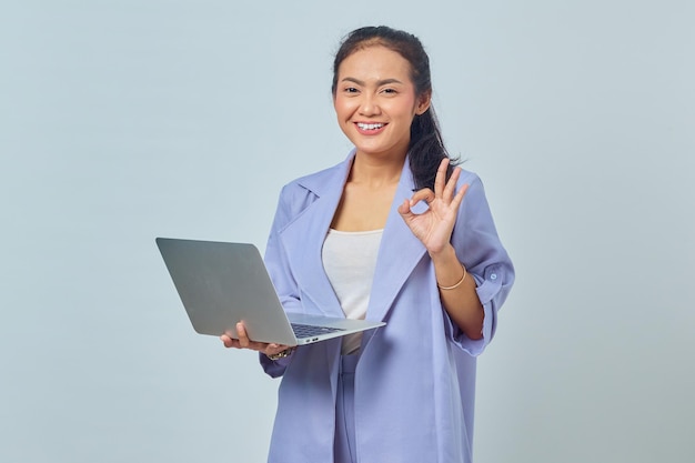 Retrato de uma jovem asiática sorridente usando laptop e gesticulando sinal de ok isolado em fundo branco