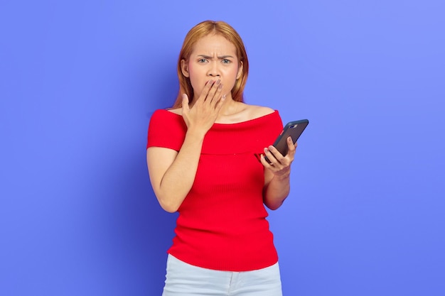 Retrato de uma jovem asiática surpresa usando telefone celular e cobrindo a boca com palmas isoladas sobre fundo roxo