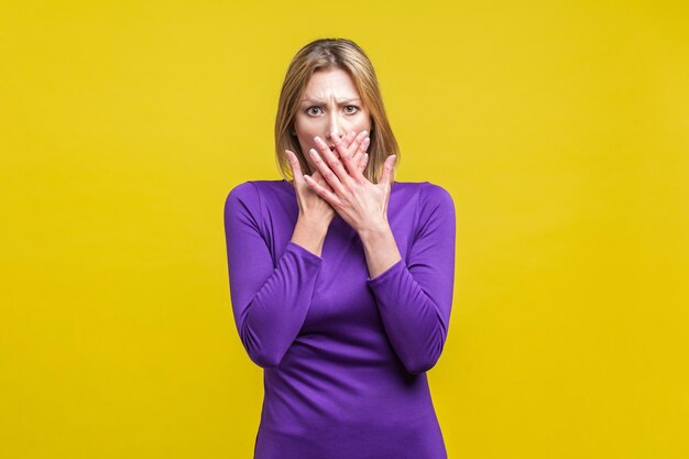 Retrato de uma jovem assustada em elegante vestido roxo apertado, cobrindo a boca com as mãos, sendo intimidada e com medo de contar sobre problemas. tiro de estúdio interno isolado em fundo amarelo