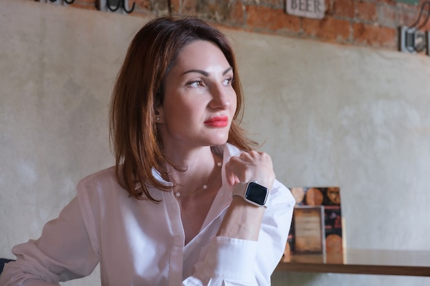 Retrato de uma jovem atraente, com cabelo ruivo na camisa branca em uma mesa de madeira em um café. A mulher descansa a cabeça com a mão e olha pensativamente para o lado.