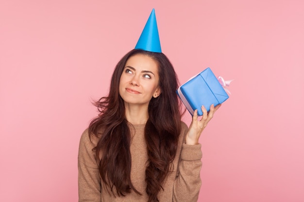 Retrato de uma jovem atraente curiosa com expressão pensativa usando cone de festa na cabeça segurando o presente e sonhando imaginando o que está dentro do estúdio de caixa tiro isolado no fundo rosa