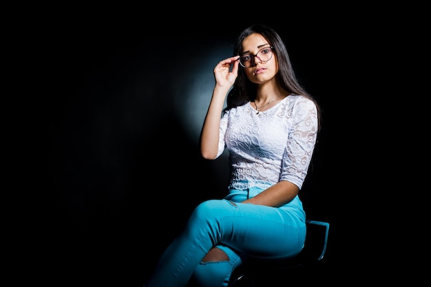 Retrato de uma jovem atraente de blusa branca e calça azul sentada posando com seus óculos no escuro