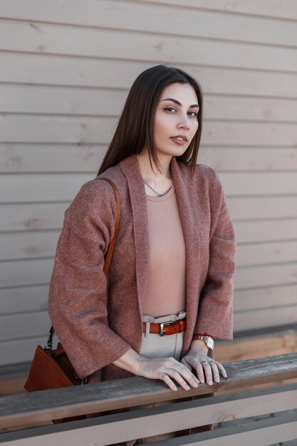 Retrato de uma jovem atraente de cabelos castanhos atraente num elegante casaco vintage com lábios sensuais perto de um edifício de madeira na cidade. Bem menina moderna em lindas roupas elegantes descansa ao ar livre no dia de primavera.