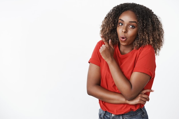 Retrato de uma jovem atraente, divertida e surpreendida, uma jovem mulher afro-americana de 25 anos, com uma t-shirt vermelha a dobrar os lábios com admiração e espanto, a apontar para o canto superior esquerdo sobre a parede branca