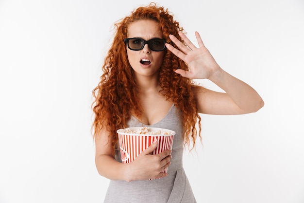 Retrato de uma jovem atraente e confusa, com cabelo ruivo longo encaracolado, isolado, assistindo a um filme e comendo pipoca