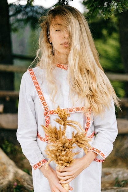 Foto retrato de uma jovem atraente loira sensível em um vestido branco com ornamento posando com buquê de espigas sobre a natureza