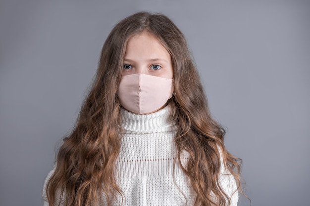 Retrato de uma jovem atraente menina com cabelos longos esvoaçantes loiros com uma camisola branca na máscara protetora médica sobre um fundo cinza do estúdio. Lugar para texto. Copie o espaço.