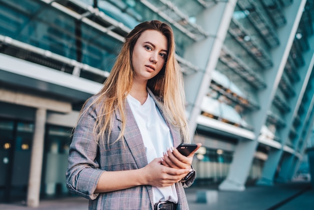 Retrato de uma jovem atraente mulher de negócios vestida casualmente com o celular nas mãos, posando em frente a um prédio de alta tecnologia