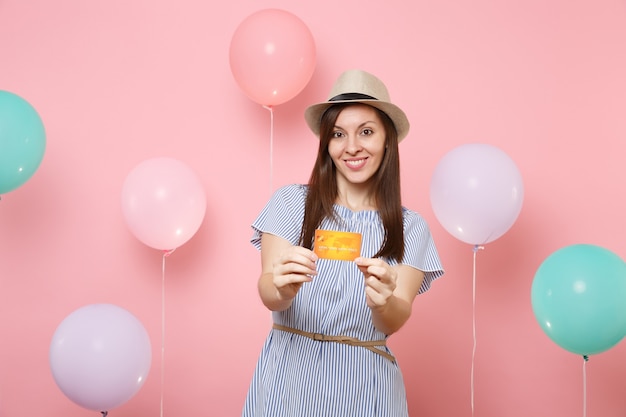 Retrato de uma jovem atraente sorridente com chapéu de palha de verão e vestido azul, segurando o cartão de crédito em fundo rosa pastel com balões de ar coloridos. emoções sinceras de pessoas de festa de férias de aniversário.