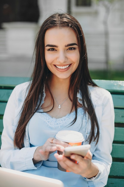 Retrato de uma jovem atraente usando um smartphone enquanto bebia café sentado em uma praia no parque com um laptop nas pernas olhando para a câmera sorrindo