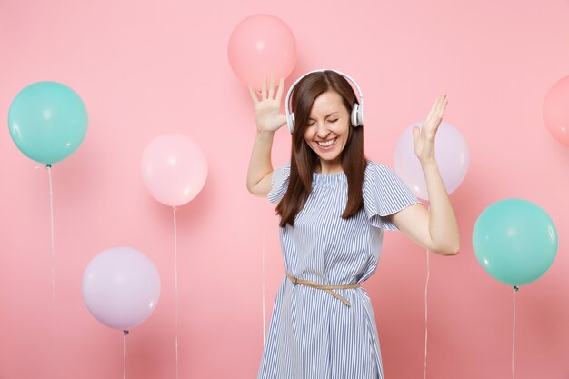 Retrato de uma jovem bonita alegre com fones de ouvido no vestido azul, ouvindo música, espalhando as mãos no fundo rosa com balões de ar coloridos. Festa de aniversário, emoções sinceras de pessoas.