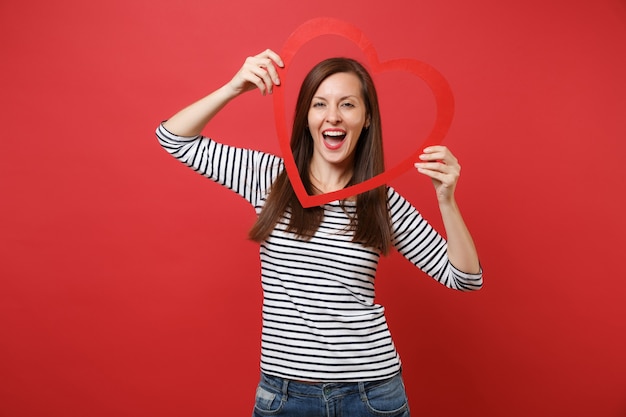 Retrato de uma jovem bonita alegre em roupas listradas casuais, segurando um grande coração de madeira vermelho isolado no fundo da parede vermelha brilhante. Emoções sinceras de pessoas, conceito de estilo de vida. Simule o espaço da cópia.