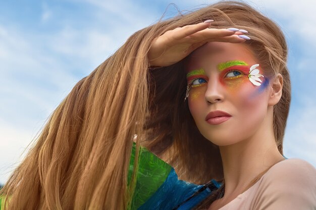 Retrato de uma jovem bonita, branca, mulher com cabelos longos e maquiagem de arte criativa, close-up em um body e uma capa de chuva, campo verde e um céu azul.