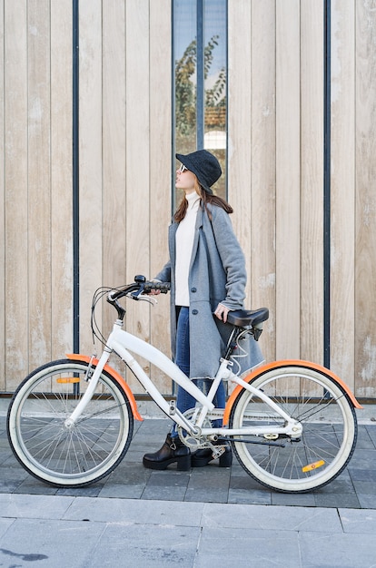 Retrato de uma jovem bonita caucasiana, vestida com uma jaqueta cinza, um suéter branco e um chapéu com um passeio de bicicleta na cidade