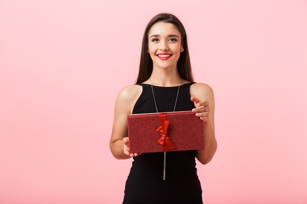 Retrato de uma jovem bonita com um vestido preto de pé isolado sobre um fundo rosa, segurando uma caixa de presente