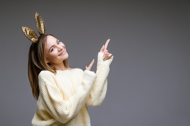 Retrato de uma jovem bonita e alegre com um suéter e orelhas de coelho mostra o dedo para o lado